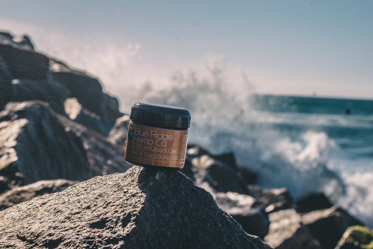 home-hero Close-up of CBD infused gel jar on rocks by the sea, showcasing product against a scenic ocean backdrop with splashing waves.